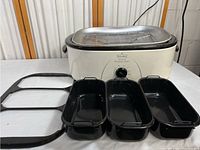 Overview of the Rival 18 Quart Roaster Oven with three black enamel roasting pans, three glass lids, and the chrome plated roasting rack beside the oven