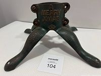 Front view of the antique Christmas tree stand showing 'A MERRY XMAS' inscription and three splayed legs on white surface.