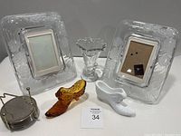 Photo showing 2 clear glass floral etched picture frames, 1 Imperial Candlewick glass vase, 1 white milk glass boot with Button and Daisy pattern, 1 amber glass boot, and small metal stand.