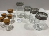 Photo showing 5 Libby glass storage jars with cork stoppers (1 large, 4 small) and 4 vintage-style glass jars with metal lids (3 Crown jars and 1 Corona jar) on white surface.