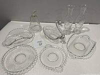 Full lot arrangement on white surface showing 2 folded fruit trays, 2 single rose vases, decorative bell, 2 handled plates, 1 plate without handles, and swan candy dish of Imperial Candlewick clear glass with beaded edges.