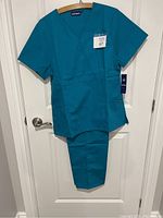 Full view of ladies teal scrub set hanging on a door, showing top and pants.