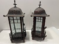 Set of two tall rectangular metal and glass lanterns with domed tops and finial handles, one hinge bent on one lantern.