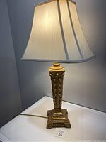 Full view of the table lamp illuminated, showing the gold ornate column base and cream fabric shade with slight marks.