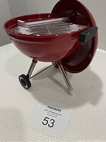 Miniature red kettle-style Weber BBQ with chrome legs, black plastic handles, and two small wheels shown from the side angle with lid off and placed behind.