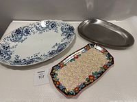 Three platters displayed on white surface: large blue and white antique porcelain platter, rectangular multicolored floral 'Rosalind' platter, and silver metal oval platter.