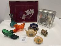Various vintage collectibles including glass boots, miniature ceramic teapot, hand-painted plate marked Made in Occupied Japan, decorative tin box, framed floral artwork, and small ceramic house figurine arranged on table.