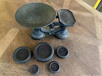 Photo showing the antique metal balance scale with a round weighing pan and a smaller rectangular platform, with six different sized weights placed in front.