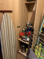 Ironing board with striped cover standing inside cabinet next to shelves holding cleaning products and a metal garbage can.