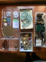 Photo showing clear plastic dresser organizer trays containing assorted small costume jewellery items and a hair clip, including pins and rings.
