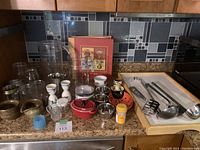 Kitchen items displayed on granite counter including glass jars, egg cups, small red casserole dish, pitchers, serving utensils, ramekins, and a recipe saver book, photographed to depict the entire lot.