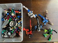 Plastic bin filled with assorted LEGO bricks and parts, as well as six partially assembled LEGO figures resembling creatures or robots with jointed limbs and claws.