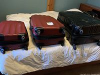 Three suitcases placed side by side on a bed, showing size and colors; two are red, one black; all have four wheels visible.