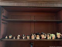 Photo showing all 12 Toby jugs arranged in a row on wooden shelves, displaying varied characters including Alice in Wonderland figures.