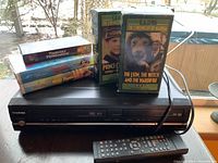 Front view of Toshiba VHS/DVD combo player, remote control, and stacked VHS tapes including classic titles.