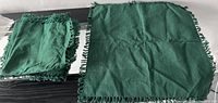 Six green cloth napkins with fringed edges, one napkin displayed flat and the others stacked.
