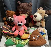 Nine assorted plush toys including bears, reindeer/moose, pink bear, doughnut, penguin, pinecone, fish tail, toy figure, and poop emoji plush arranged on grey fabric background