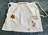 Front full view of Bancroft rock hound paradise white apron with embroidered rock hound figure, text, and geometric shapes.