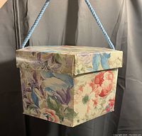 Full view of the floral hat box showing the blue rope handle and square shape with lid.