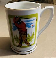 White ceramic mug with colored golfer illustration and gold trim.