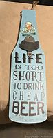 Light blue wooden hanging bottle opener shaped like a paddle with a black metal bottle opener and beer-themed text and graphic.