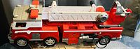 Side view of large toy fire truck showing the full length with extendable white ladder, red and white color scheme, multiple chrome detailed wheels, and attached fireman figurine on the cab roof.
