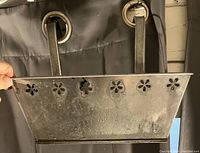 Photo showing rectangular metal planter with floral cutout along the top edge and two metal hooks for hanging. Surface shows wear and oxidation.