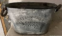 Oval galvanized metal planter with rope handles and decorative embossed text 'FLOWERS & GARDEN' on front.
