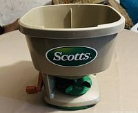 Photo of Scotts seed spreader showing green and beige plastic hopper with Scotts logo, handle, and adjustable dial.