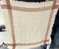 Square woven cotton tablecloth showing its red patterned border and fringed edges against a black background held up by a person.