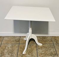 White wooden accent table with rectangular top and a three-legged pedestal base, standing on tiled floor against a white wall.