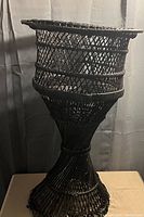 Full side view of black woven wicker floor model plant stand showing flared base and basket top.