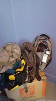 Four bags on a box against a blue wall including REI backpack, brown travel bag, Under Armour backpack with tags, and black/yellow sports bag.