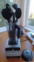 Vita-Mix blender with clear container holding black cooking utensils on wooden table near window