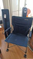 Full view of one blue MacCabee folding camp chair in opened position with its carry bag standing upright behind it.