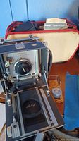 Opened Linhof Technika camera showing black bellows, front standard with Wollensak Rapax shutter lens, Linhof lens cap on film bed, leather-cased light meter, blue cloth, red-trimmed cream Polaroid case in background