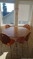 Full view of round dinette table with four matching chairs around it. Wooden finish with metal legs, located in a sunlit room by windows.