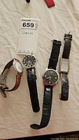 Four wristwatches laid out on fabric surface with auction tag showing lot number 659. Two watches have black leather straps, one has brown leather strap, one is rectangular faced.