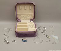 Photo showing purple velvet zippered travel storage box with three open compartments and several silver chains, pendants, ring, and stud earrings laid out in front.