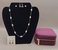 Photo showing pearl necklace on black display bust, various pairs of earrings on cards, and purple zippered travel storage box.