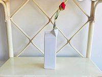 Front view of white ceramic milk box vase with a single red artificial flower inside, placed on a chair with a woven back.