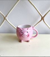 Front-facing view of the pink ceramic pig mug with a detailed pig face and feet visible, placed on a light-colored surface with a white rattan background.