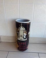 Front angle showing the cylindrical ceramic umbrella holder with vintage cafe artwork, black background, and red text.