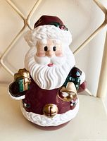 Front perspective showing detailed Santa Claus face, hat, and the hand holding a bell and gift.