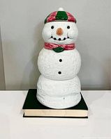Front view of white ceramic snowman cookie jar with red and green hat and red scarf atop a book