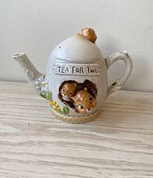 Front and side view of the cream-colored ceramic teapot with floral and puppy decorations and embossed phrase 'TEA FOR TWO'.