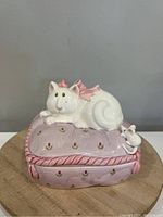 Front view of vintage ceramic cat storage box showing white cat with pink wings on lavender cushion with pink trim and small floral motifs, mouse figure beside cat.