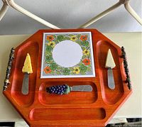 Top view of vintage wooden cheese tray with decorative ceramic tile and three cheese spreaders with themed handles.