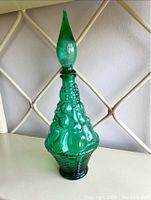 Full view of green glass decanter with textured geometric pattern and matching pointed stopper, photographed on a chair back.