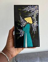 Closed view of the rectangular black jewelry box with hand-painted design of a figure in a teal outfit and yellow hat surrounded by purple flowers and green leaves.